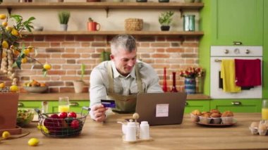 Good looking man in the morning doing a online shopping while standing at the kitchen island using the credit card and laptop pretty woman hugging with love her husband. 4k