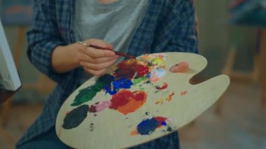 A close up shot of the painter woman dabbing into her collide palette with her brush and painting something onto the canvas while she vibes with the music. Shot on ARRI Alexa