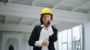 In a large space construction site beautiful woman architect speaking in her smartphone and looking around at the other workers.