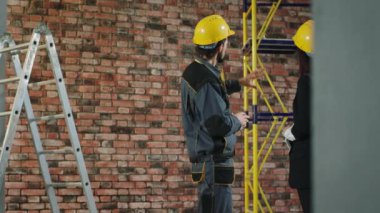 Two focused woman and man builders in blue coveralls discussing workflow while standing at construction site indoors with papers in hands.