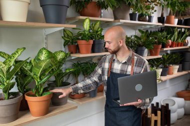 Çiçekçi dükkanındaki beyaz girişimci dükkan envanterini kontrol etmek için dizüstü bilgisayarını kullanıyor sonra da kameraya bakıp gülümsüyor..