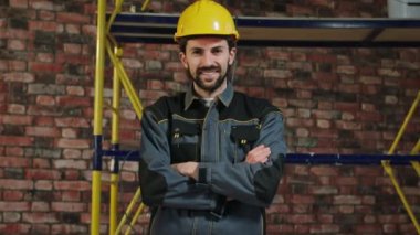 Charismatic with a large smile constructor worker posing in front of the camera crossing hands he wearing safety helmet and equipment. 4K