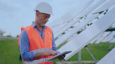 Güvenlik üniformalı karizmatik olgun bir ekoloji mühendisi. Kameraya yakın. Fotovoltaik güneş panellerinin verimliliğini ve dijital tablet yapmak için elinde tuttuğu güneş operasyonunu analiz ediyor.