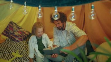 Good looking boy with long hai together with his daddy reading a story book together in the tent at home. 4k