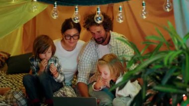 In front of the camera capturing video of young family with two cute kids watching movie together in the kids room inside the handmade tent.