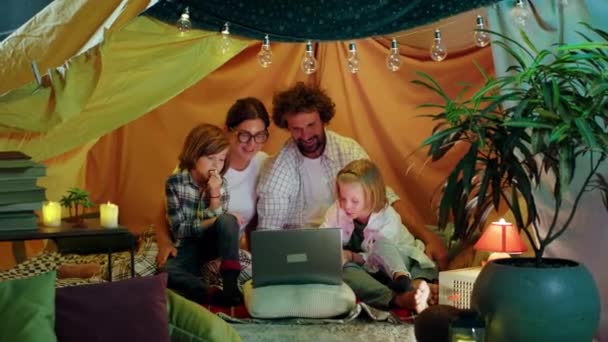 かわいい若い家族の両親と2人のかわいい子供たちが一緒に時間を楽しんでいる床の上の子供部屋の余分なテントの中に座っている間に映画を見ているのは幸せな時間です — ストック動画