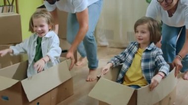 In front of the camera after young family moving to new home they take their kids and put them on the boxes and start to doing a ride through the living room. 4k