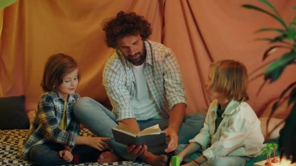 彼らはすべての多くの枕に囲まれた毛布屋内テントで本を読んでいるので 非常に幸せなお父さんは彼の2人の小さな男の子と楽しい時間を過ごしています — ストック動画