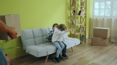 Genç bir aile yeni bir eve taşınıyor mutlu çocuklar oturma odasındaki kanepede otururken ve aileleri son kutuları taşırken büyük bir gülümsemeyle eğleniyorlar. 4k