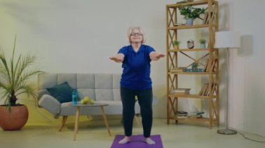 Evde, kameranın önünde gülümseyen, tatlı yaşlı kadın antrenmanı yaparken sağlıklı bir vücut için egzersiz yapıyor yoga minderinin üzerinde duruyor ve kameranın önünde egzersiz yaptıktan sonra poz veriyor.