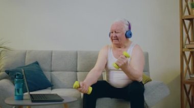 Evde, oturma odasında, karizmatik yaşlı bir adam dizüstü bilgisayara bakarken ve kulaklıkla egzersiz yaparken koltukta oturur ve pazıları ağırlık ile eğitir. 4k