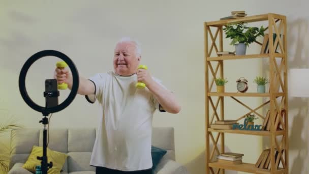 自宅のリビングルームでカリスマ老人は非常に自宅で彼の運動のビデオを記録するためにリングライトを使用して幸せ彼は運動を行うための重みを使用しています — ストック動画