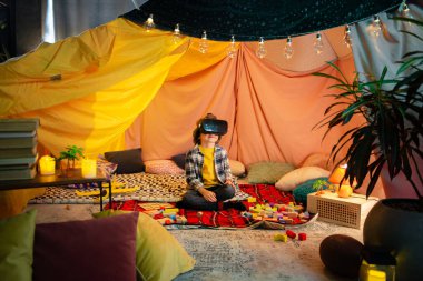 One little blond boy is showing things to another brunette boy who is wearing a vr headset inside a comfortable sheet fort.
