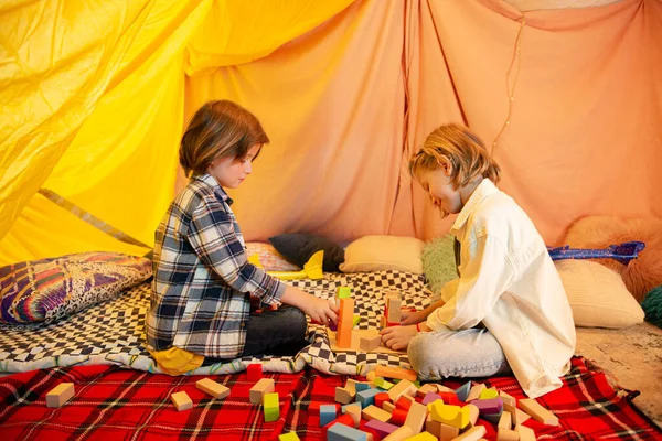 Two young boys are messing around with toy airplanes inside a big blanket tent which has very bright lighting and is decorated in a very modern way to create a comfortable atmosphere. Portrait