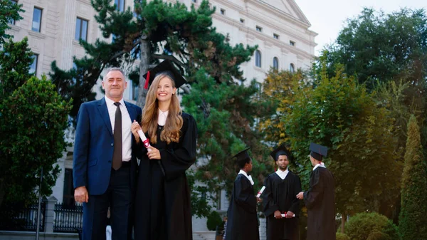 大学の庭で美しい女性の学生のポーズ興奮彼女の卒業証書とともに大学教授カメラの前で — ストック写真