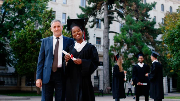 卒業日カリスマ黒人女性卒業生が卒業証書を保持しながらカメラの前で大学教授とポーズ — ストック写真
