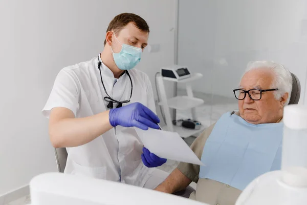 在牙医面前 有魅力的牙医向他的病人解释植入物是如何固定在牙医椅子上听的 — 图库照片