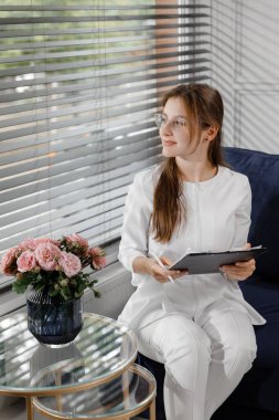Tıp kliniğinde kameranın önünde mutlu ve güzel doktor kadın terapist pozu veriyor. Kanepede oturuyor ve tatlı tatlı gülümsüyor. Hastane