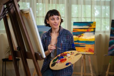 Yaratıcı genç kadın ressam projesi için yeni bir sanat çalışması üzerinde çalışıyor ve resim stüdyosunda poz veriyor. Resimlerinin önünde duran neşeli bir ressam. Boya