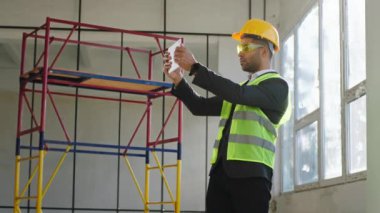 İnşaatın fotoğraflarını çekmek için dijital tablet kullanan yakışıklı bir işadamı koruyucu ekipman ve gözlük taktığı sonucunu analiz ediyor..