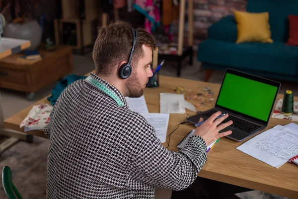 Happy and charismatic designer guy at his office in front of the camera have a conversation with a customer he speaks online from the laptop using the headphones the desktop of laptop are green