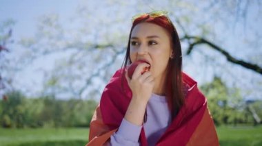Parkın ortasında, elmayı yiyen ve güneşin tadını çıkaran ve lgbt bayrağını tutan güzel bir lezbiyen kadın. 4k