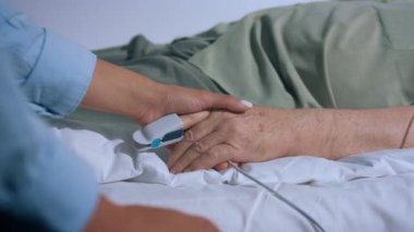 Hastanedeki yaşlı hasta ameliyattan önce yatağa uzanmış ve parmak kalp ritmi takıyormuş. Aile ferdi kadın onu el ele tutuşmaya teşvik etmiş. 4k