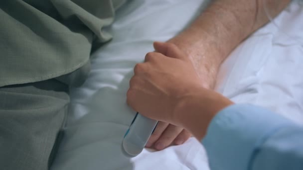 ベッドの上に寝そべっている老人の病室では 娘が脆弱な手を握っている間に回復し休息している手に焦点を当て サポートと慰めを与えています — ストック動画