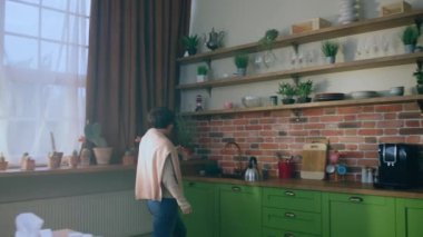 Sabah kısa saçlı bayan lavabodan su ısıtıcısını alır ve ısıtıcıyı elektrikli ocağa koymak için mutfak adasına doğru yürür. 4k