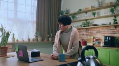 Kısa saçlı, karizmatik bir kadın mutfaktaki adanın yanında su kaynamasını beklerken dizüstü bilgisayara bakıyor. 4k