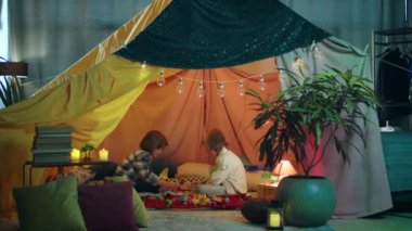 İki genç çocuk büyük renkli ve canlı bir battaniye çadırının içinde kıpırdanıp uçaklarla oynuyorlar..