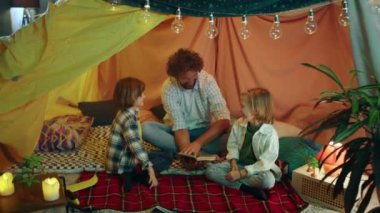 Bir baba çocuklarıyla birlikte büyük ve geniş bir battaniye çadırında onlara kitap okurken ve birlikte geçirdikleri zamanın tadını çıkarırken gülüyor. 4k