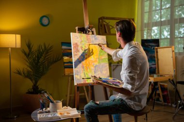 Sanat stüdyosunda yakışıklı bir sanatçı boya fırçasını alır ve sandalyede otururken tuvale yeni bir resim çizmeye başlar. Boya