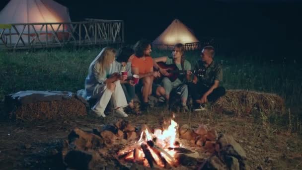 友人のカリスマ的かつ魅力的な幸せなグループは 火災の杭の横にある干し草の上に座っている間 彼らはギターで演奏を歌うキャンプ場で楽しい時間を過ごす Arriで撃たれた — ストック動画