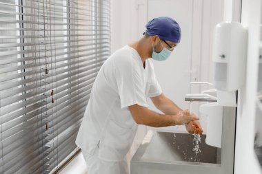 Ameliyat odasındaki modern hastanede ameliyata başlamadan önce doktor cerrahın önünde ellerini iyice yıkamak ve tıbbi üniforma giymek ve koruyucu maske takmak. Hastane