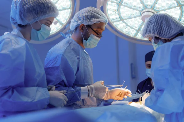 Three medical surgeons in scrubs and one standing at the head of the patient in dark blue scrubs are operating a surgery and talking.