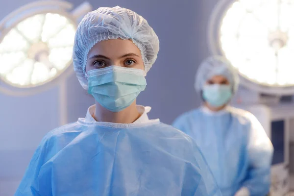 一个女护士的近照 她直接看着摄像机 看上去很美 而在她身后站着一个医疗大灯和一个护士 — 图库照片