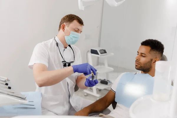 在一个现代的牙科诊所里 戴着保护膜的牙医在牙齿模型上向病人解释牙齿上的支撑是如何看待坐在牙医椅子上的病人的 牙科医生 — 图库照片