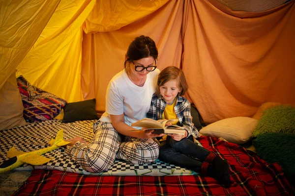 A brunette pretty young mother is bonding with her blonde little boy while reading him a bedtime story and chilling in a large snug indoor tent. Family