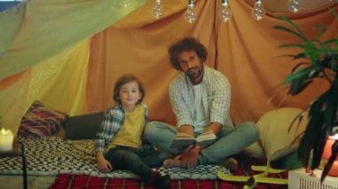 Concept of happy childhood dad and his cute son posing in front of the camera while speeding good time together on the tent in the kids room. 4k