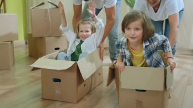 In the large modern living room happy kids have a ride on the boxes while their parents carrying them through the all living room in the new house. 4k