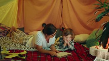 A mother wearing comfortable pyjamas is laying down next to her son and is reading out a book to him in this very wholesome moment they are also inside a modern and large indoor tent. 4k