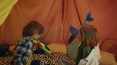 Two youthful boys are playing joyfully together with airplanes and making them crash into each other all while being in a big blanket tent.