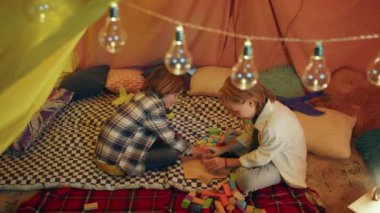 Two junior boys are engaging in building blocks together while sitting inside a multicolored sheet tent.