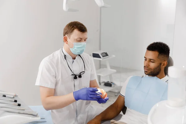 保護マスクを持つ良い外見の男性歯科医は 患者に彼が示すと簡単に説明するために歯モデルを使用して仕事のプロセスを説明します — ストック写真