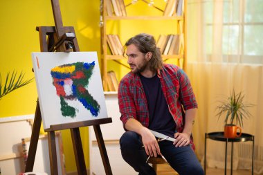 Orta saçlı ve sakallı yakışıklı bir ressam sanat eserine bakıyor ve fırçası ve paletiyle üzerine resim yapıyor..
