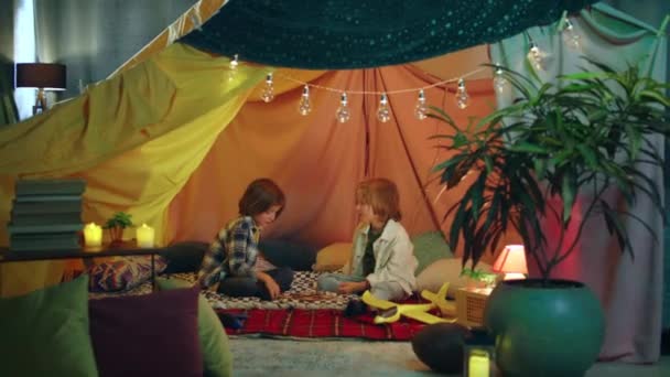 二人の若いかわいい男の子がそれにヒッピーの雰囲気を持っている素敵な快適なテントの中で遊んでいます テントは屋内のものです — ストック動画