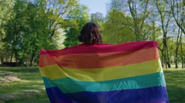 Parkta güneşli bir gün. Kısa saçlı lezbiyen. Elinde lgbt bayrağı, çimenlerde yürüyor..