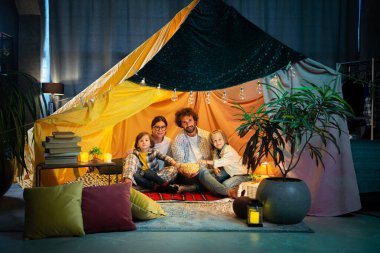 In the kids room down on the floor parents and their kids watching a movie while eating popcorn they sitting inside of handmade tent.