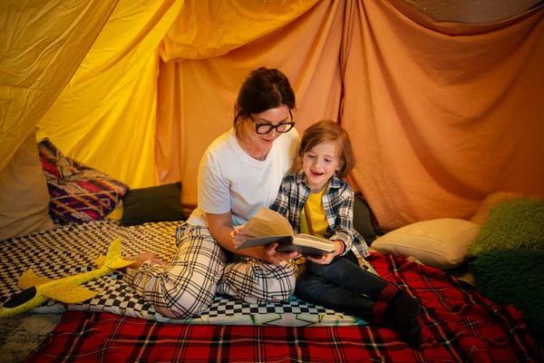 A brunette pretty young mother is bonding with her blonde little boy while reading him a bedtime story and chilling in a large snug indoor tent.n bFamily